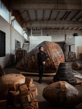 Porträt einer schwarz gekleideten Künstlerin, die in ihrem Atelier an einer großen Holzskulptur lehnt und nach oben schaut. 