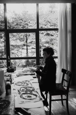 Redaktionelles Porträt in Schwarzweiß einer Künstlerin, die an ihrem Tisch sitzt und in einem Skizzenbuch blättert. 