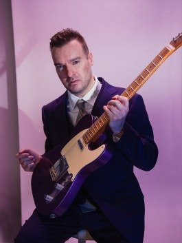 Porträt eines Gitarristen, der mit der Gitarre in der Hand, lila Outfit und rosa Hintergrund sitzt und zur Fotografin blickt. 